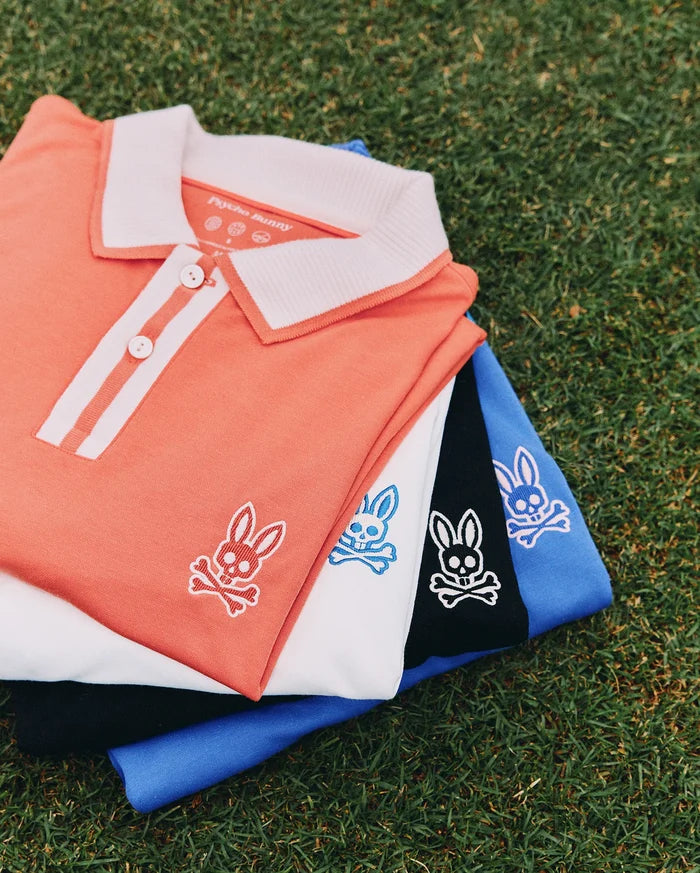 Mens Ridley Pique Polo stack: coral with white ribbed collar and placket stripes, white, black, and royal blue pique polos, each featuring a small embroidered Psycho Bunny skull logo on the left chest.