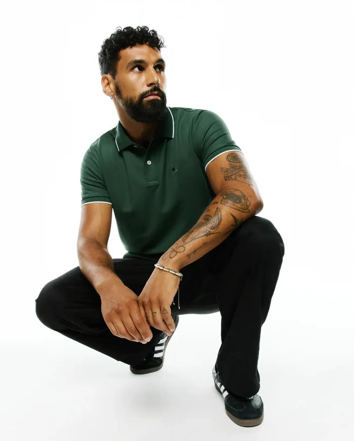 Mens Ringston Pique Polo in dark green, featuring a classic fit, pique knit fabric, white tipping on the collar and cuffs, and a small embroidered Psycho Bunny skull logo on the left chest.