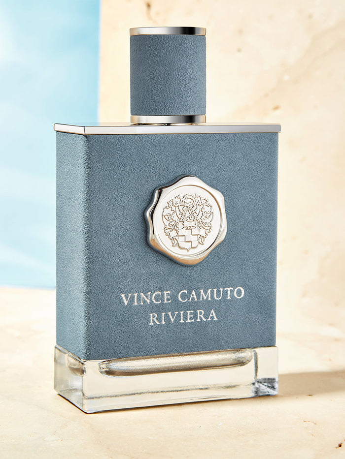 A blue and silver bottle of Vince Camuto Men’s Riviera 3.4 oz Eau de Toilette Spray rests on a light stone surface against a blue and beige background, evoking Mediterranean energy with notes of Italian lemon and lavender oil.