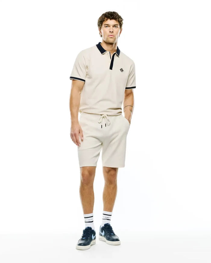 Mens Robin Pique Polo in cream with navy collar and cuff tipping, featuring a small embroidered Psycho Bunny skull logo on the left chest, paired with matching cream drawstring shorts.