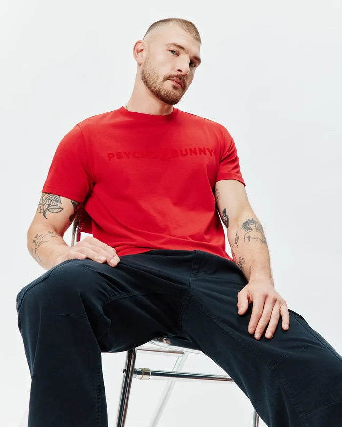 Mens Royce Graphic Tee in vibrant red, crafted from soft jersey knit with a classic fit, featuring a large, tonal embossed PSYCHO BUNNY brand name across the chest, complemented by dark blue cargo pants.