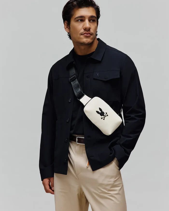 Mens Rubberized Crossbody in cream with a large black Psycho Bunny skull logo, worn with a black collared shirt jacket featuring a small embroidered bunny, a black crew neck tee, and khaki pants.