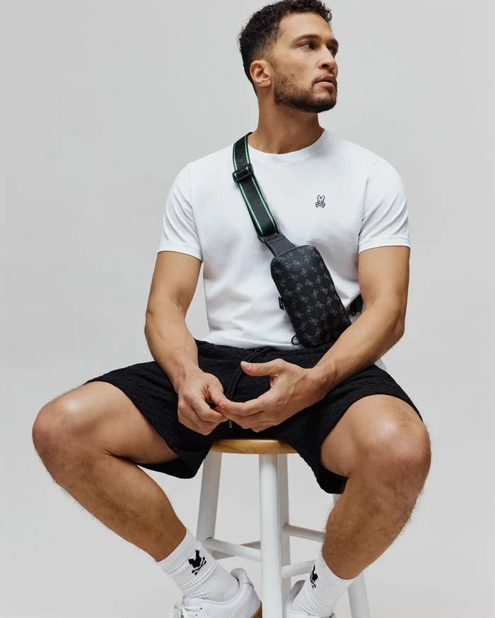 White jersey t-shirt with embroidered pink-eared Psycho Bunny skull, black textured shorts, and a black Mens Rubberized Crossbody bag with all-over bunny skull print and green-striped strap.