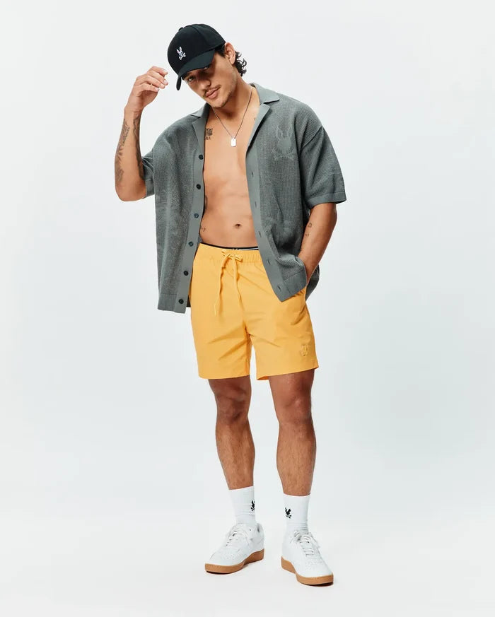 Mens Samos Hydrochromic Printed Swim Trunk in bright yellow with an embroidered Psycho Bunny skull logo, a relaxed charcoal knit short-sleeve shirt with subtle skull pattern, and a black cap with an embroidered skull.