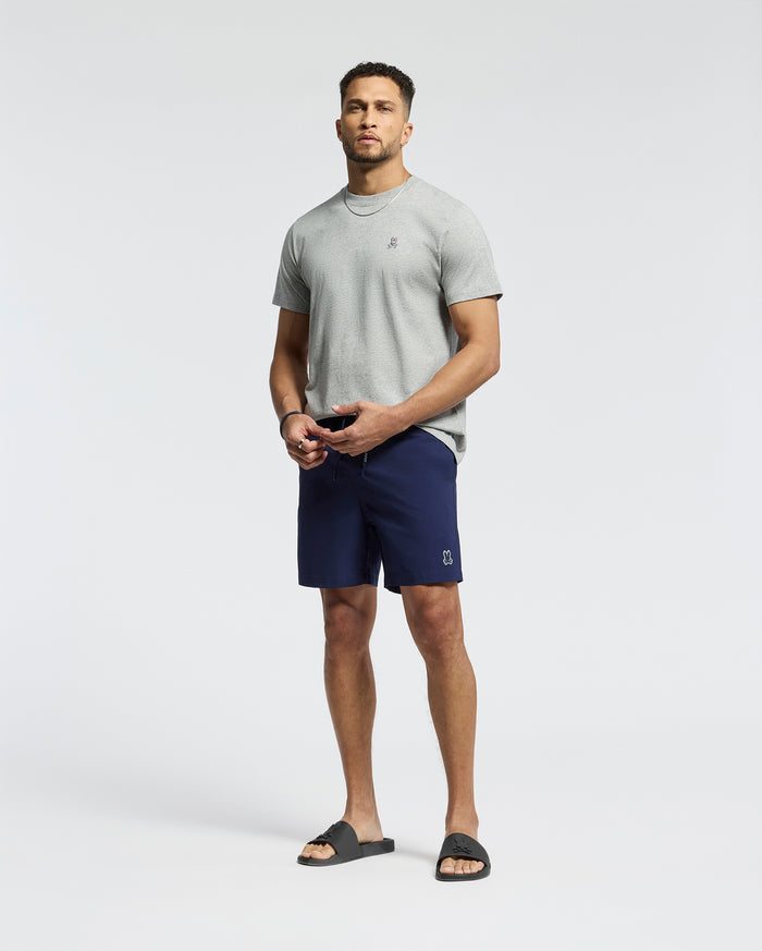 A man stands against a plain background wearing a light gray T-shirt and Psycho Bunny MENS SAMOS HYDROCHROMIC PRINTED SWIM TRUNK. He completes the look with black slide sandals, has a short beard, and gazes at the camera neutrally.