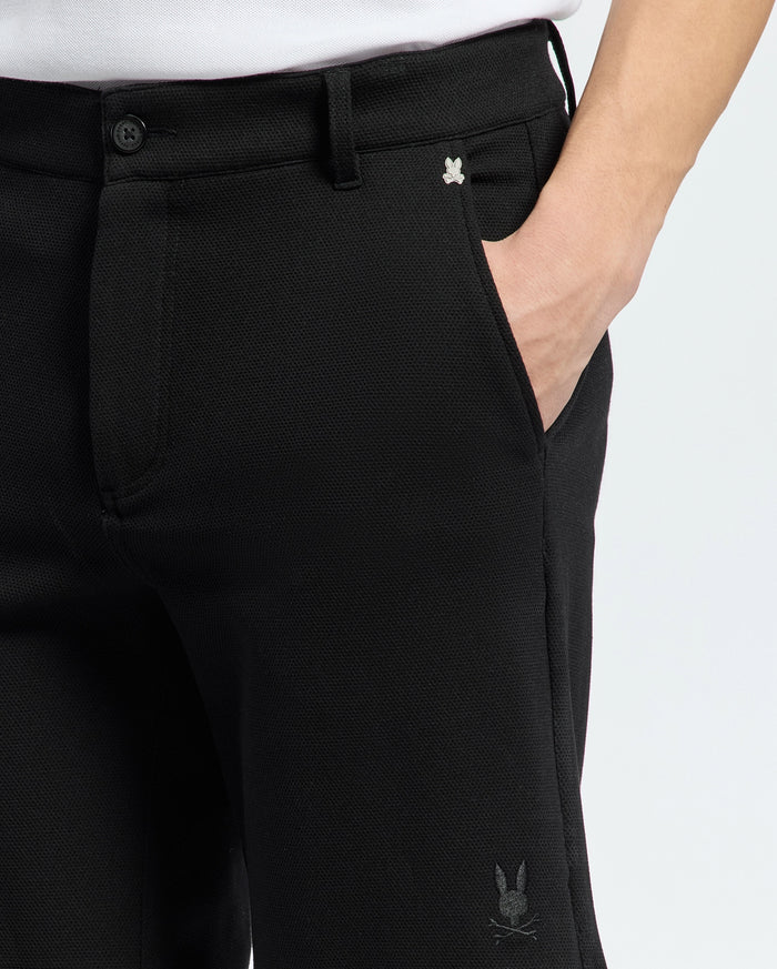 A person wears MENS SHIRO KNIT HONEYCOMB SHORT by Psycho Bunny. Black shorts feature a small embroidered rabbit logo near the pocket and on the leg, showcasing chic simplicity against a plain white background.