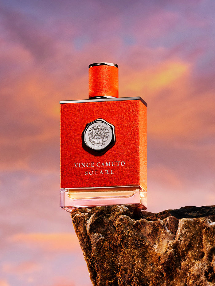 A bold orange bottle of Vince Camuto Men’s Solare 1.7 oz Eau de Toilette Spray rests on a textured rock, set against a vivid sunset sky, perfectly reflecting the dynamic spirit of this woody oriental men’s fragrance.