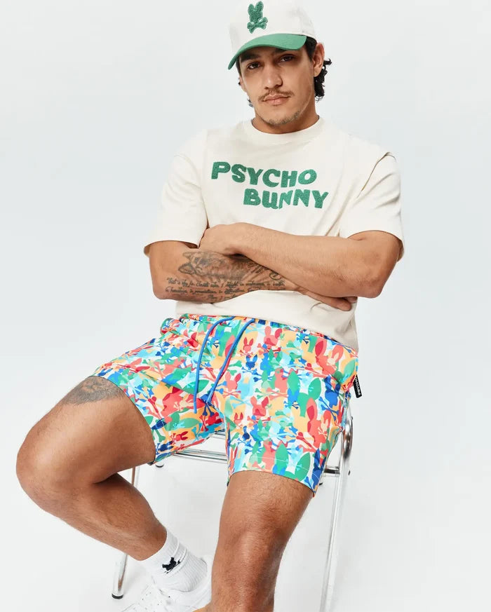 Mens Tanner Swim Trunk in vibrant multi-color with all-over Psycho Bunny skull print and blue drawstring. Styled with a cream jersey t-shirt featuring bold green PSYCHO BUNNY text and a two-tone cap with bunny logo.