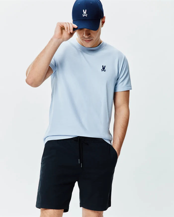 Light blue Mens Taylor Pique Tee with a small navy embroidered Psycho Bunny skull logo on the left chest, paired with dark navy drawstring shorts and a navy cap featuring a white and pink embroidered bunny skull.