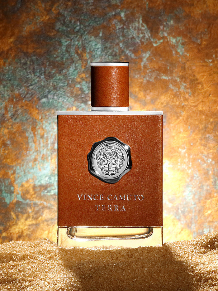 A bottle from the Vince Camuto Men’s Terra 2-Piece Set stands upright in sand, showcasing a brown leather-like covering and a metallic emblem, set against a textured, illuminated background. This refined fragrance captivates with bold notes of Sicilian bergamot blended with modern woods, creating an unforgettable masculine scent experience.