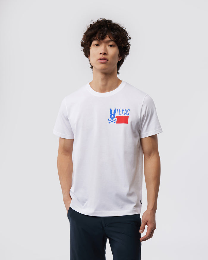 A young man with curly hair wearing a white MENS TEXAS TEE - B6U553W1PC shirt with a "Psycho Bunny" logo on the chest, paired with dark pants, standing against a plain background.