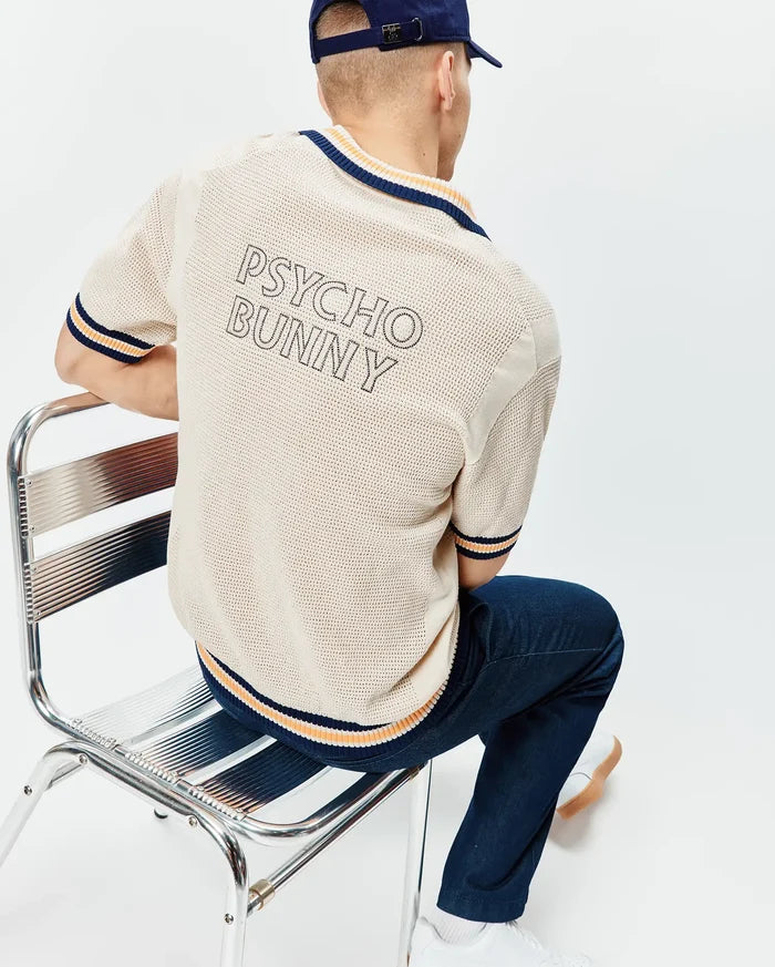 Cream Mens Theros Knit Polo with a relaxed fit, featuring an open knit texture, navy and orange striped ribbed collar, cuffs, and hem, and a large outlined PSYCHO BUNNY text logo on the upper back.