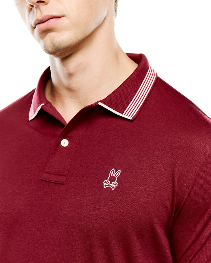 Mens Timmy Interlock Polo in deep maroon, featuring a smooth interlock knit fabric, classic fit, ribbed collar with white tipping and three white stripes, and a small white embroidered Psycho Bunny skull logo on the left chest.