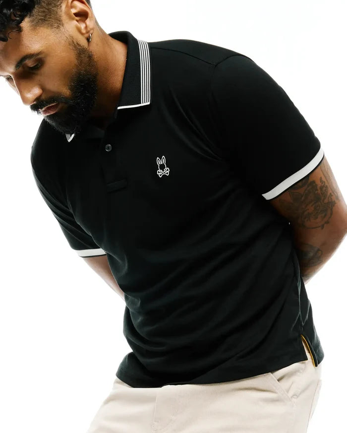 Mens Timmy Interlock Polo in black, featuring a classic fit, interlock fabric, white striped collar, white ribbed cuffs, and a small embroidered white Psycho Bunny skull logo on the left chest, with yellow side vent lining.