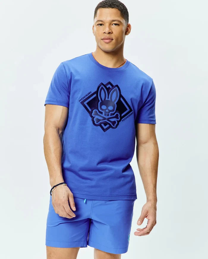 Mens Tobias Graphic Tee, a classic fit royal blue jersey t-shirt with a large dark blue Psycho Bunny skull and crossbones logo graphic in a diamond on the chest, and subtle orange collar trim.
