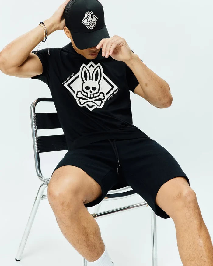 Black Mens Tobias Graphic Tee, classic jersey fit, with a large white Psycho Bunny skull and crossbones diamond chest graphic. Paired with black textured drawstring shorts and a black mesh cap with white logo.