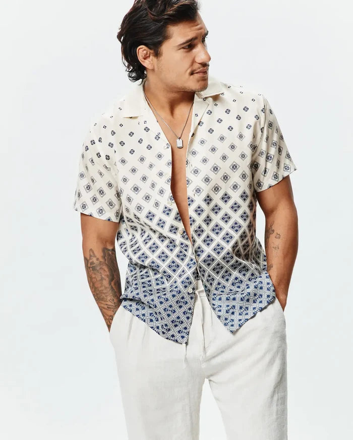 Mens Tobias Tencel Shirt, a relaxed fit cream short-sleeve shirt featuring a navy blue geometric pattern with integrated Psycho Bunny skull logos, transitioning to a denser pattern at the hem.