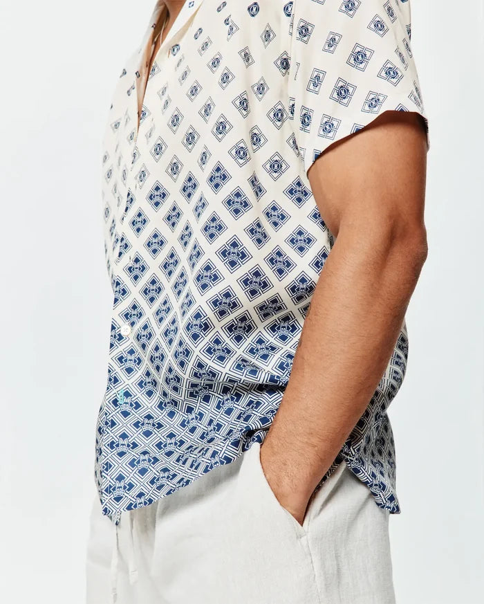 Mens Tobias Tencel Shirt, a relaxed fit cream short-sleeve button-up with a navy blue geometric and Psycho Bunny skull logo all-over print, featuring a subtle gradient effect and a small green hem tag.