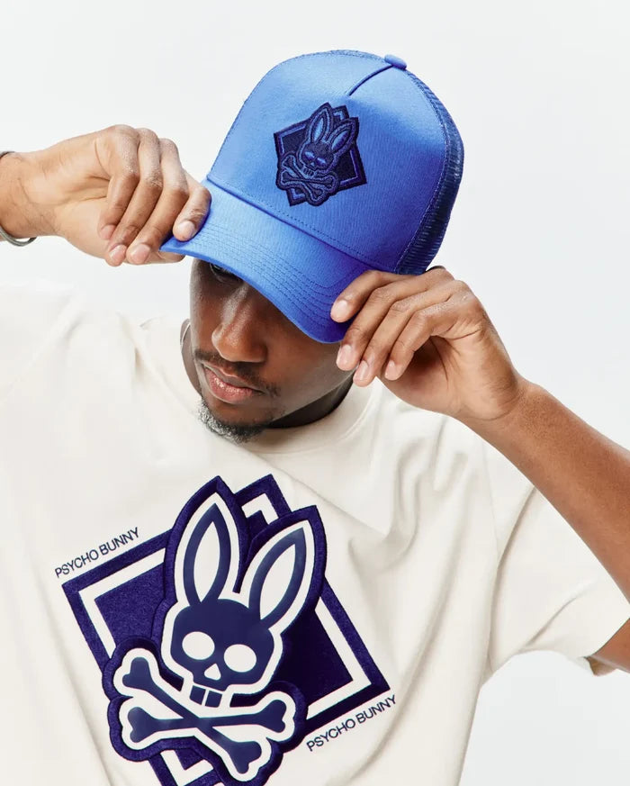 Off-white jersey t-shirt with a large central layered dark blue and white Psycho Bunny skull logo, and a bright blue mesh Mens Tobias Trucker Cap with a dark blue embroidered bunny skull patch.