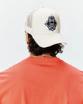 Mens Tobias Trucker Cap in cream with a prominent navy and black embroidered Psycho Bunny skull logo within a diamond on the front, featuring a mesh back. Paired with a coral jersey knit t-shirt.