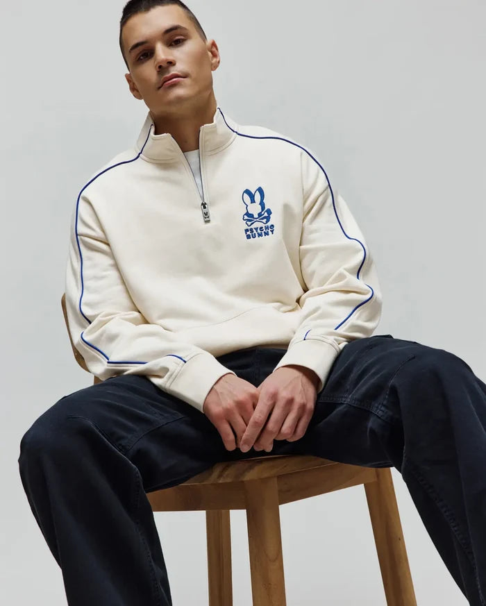 Mens Toby 1/4 Zip, a cream relaxed fit French terry sweatshirt with royal blue piping on sleeves and collar, featuring an embroidered royal blue Psycho Bunny skull logo on the left chest and a branded zipper pull.