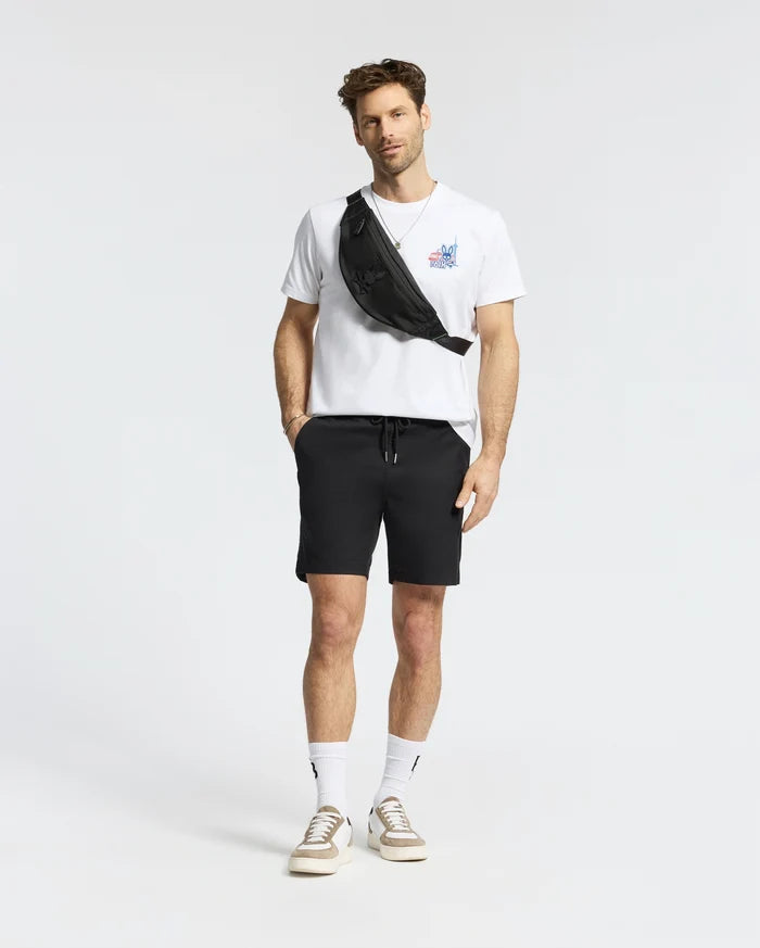 A man stands against a plain background wearing a white Psycho Bunny MENS TORONTO TEE (B6U383F200), black shorts, white socks, beige sneakers, and a black crossbody bag.