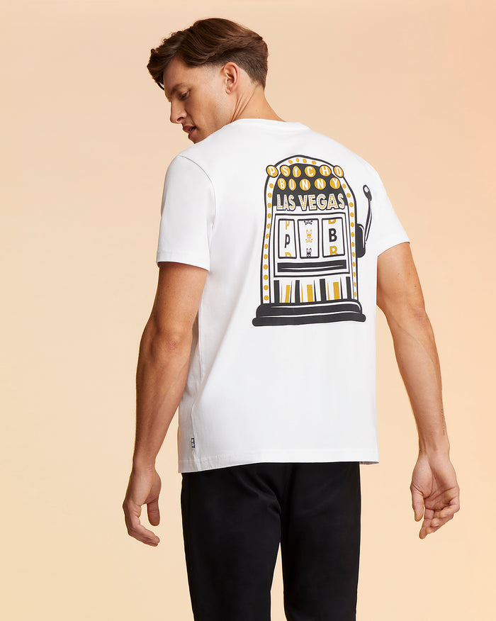 A man with brown hair is facing away from the camera, wearing a white MENS VEGAS CITY TEE - B6U967X1PC by Psycho Bunny with a colorful graphic of a Las Vegas-themed slot machine on the back. The background is a gradient of peach to beige tones. He is also wearing black pants.