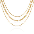 Four gold chains from the Vince Camuto Multi Layered Chain Necklace, featuring varied link styles—including a sleek snake chain and an elegant wheat chain—hang side by side on a white background to create a stylish layered look. Each chain showcases unique thicknesses and designs, lending a sophisticated, modern appeal.
