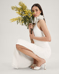 A woman in a white sleeveless dress and Vince Camuto Paigely Mule sandals crouches, holding yellow flowers and wearing a soft white ruffle accessory at her neck, set against a plain light background.