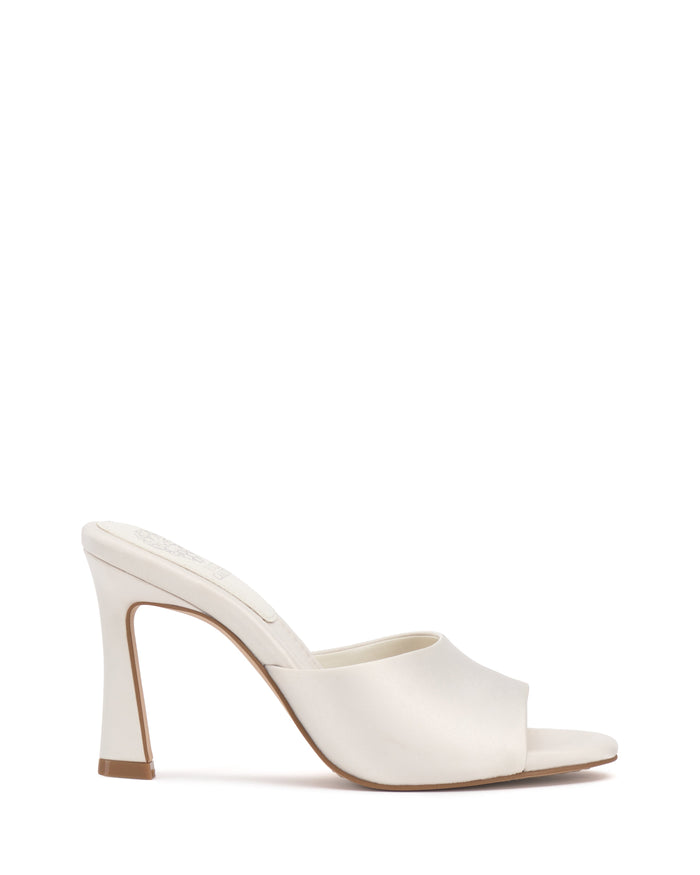 The Vince Camuto Paigely Mule is a white, open-toe satin mule with a sleek satin finish and a curved, flared heel. Featuring a luxurious pillow footbed for added comfort, it is pictured from the side on a white background.