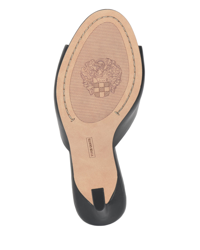 The Paigely Mule by Vince Camuto features a black minimalist design with a tan sole, accented by a crest-like motif and stitched detailing around the edge.