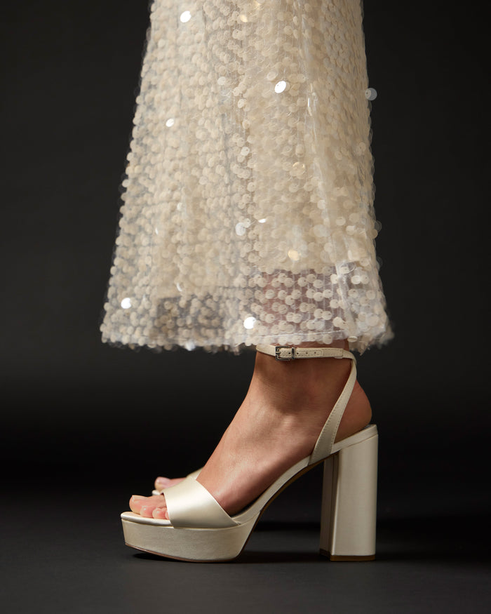 A close-up side view of a person wearing the Vince Camuto Pendry Platform Sandal in cream with block heels and a long, sheer sequin dress, set against a dark background for a chic retro vibe.