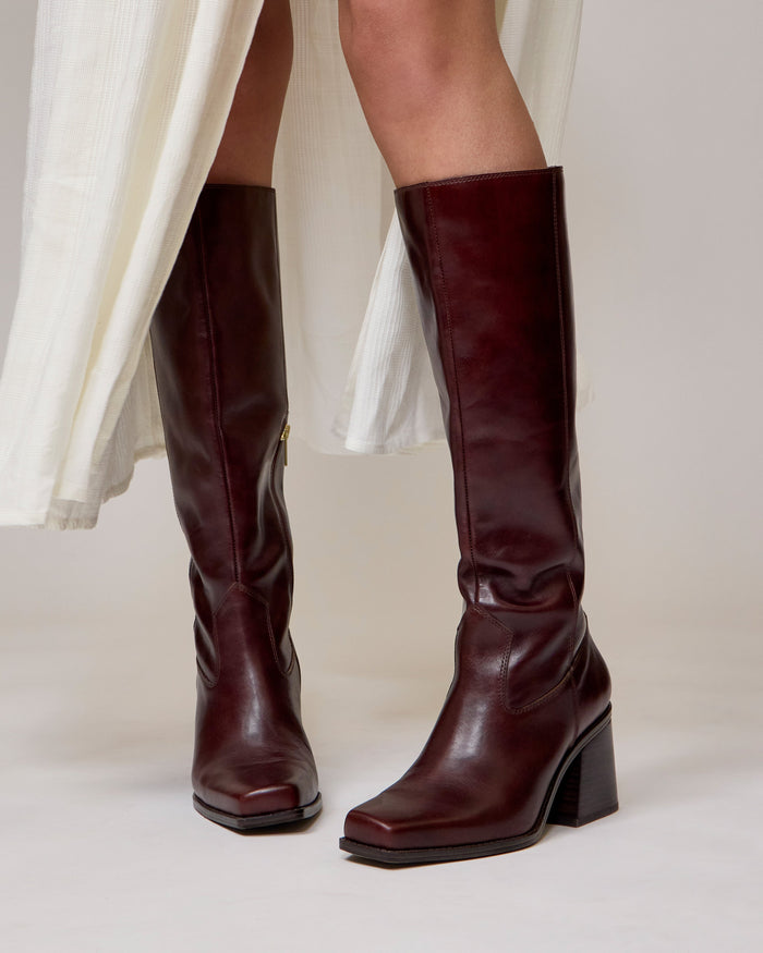 A person in a white skirt wears Vince Camuto Sangeti Boots—dark brown, knee-high, with block heels—standing on a light-colored floor.