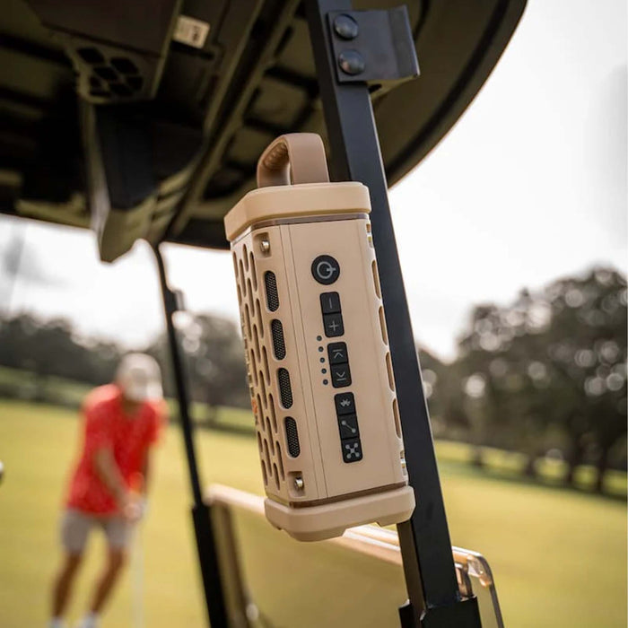 A tan Turtlebox Ranger speaker magnetically mounted to a golf cart support bar, with a golfer putting on the green in the background.