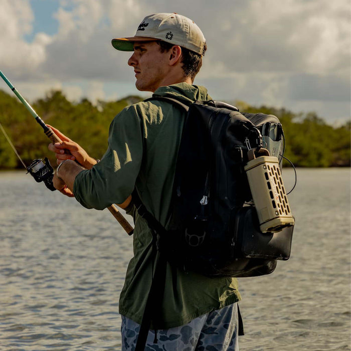 A man stands in shallow water holding a fishing rod with a black backpack that has a tan Turtlebox Ranger speaker clipped to the side.