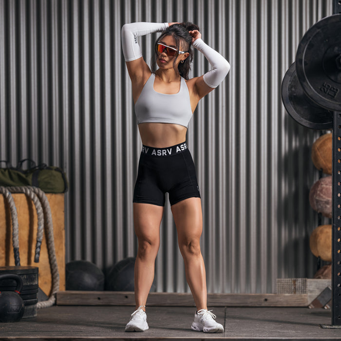 Black Lycra ribbed shorts worn with white sports bra and arm sleeves, model posing against corrugated metal backdrop