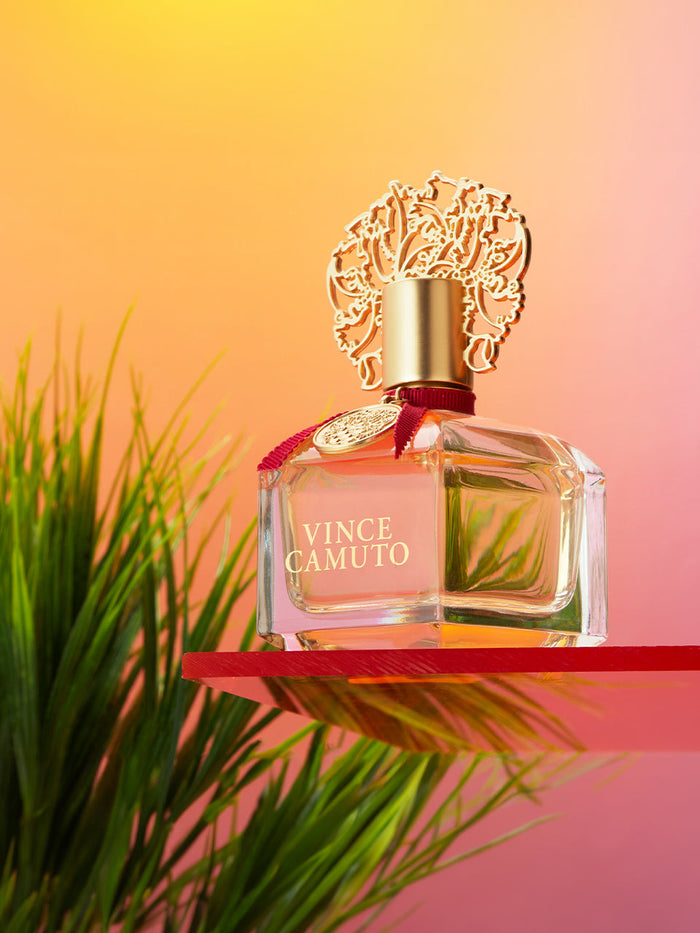A bottle of Vince Camuto Women 1.0 oz Eau de Parfum Spray, adorned with a gold ornate cap, rests on a red surface with lush green grass in the background and a warm orange-pink gradient backdrop. The luxurious scene reflects the elegant blend of Bulgarian rose and subtle leather notes that characterize this signature fragrance by Vince Camuto.