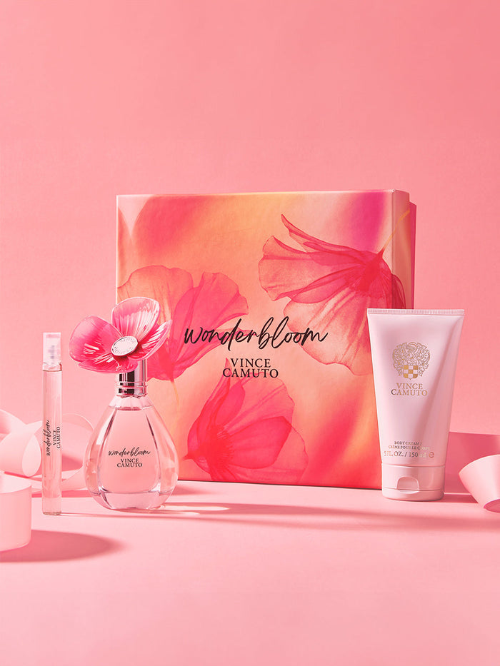 The Vince Camuto Wonderbloom 3-Piece Set is shown on a pink background with a floral perfume bottle, body cream tube, travel spray, and a decorative box featuring pink and orange flower designs.