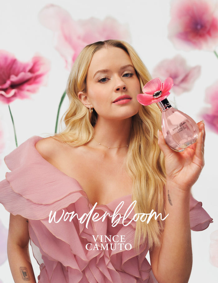A woman in a pink ruffled dress holds the Vince Camuto Wonderbloom 3-Piece Set, featuring a floral-capped bottle, surrounded by soft pink blooms and sandalwood accents on a white backdrop. Text reads: Wonderbloom 3-Piece Set VINCE CAMUTO.